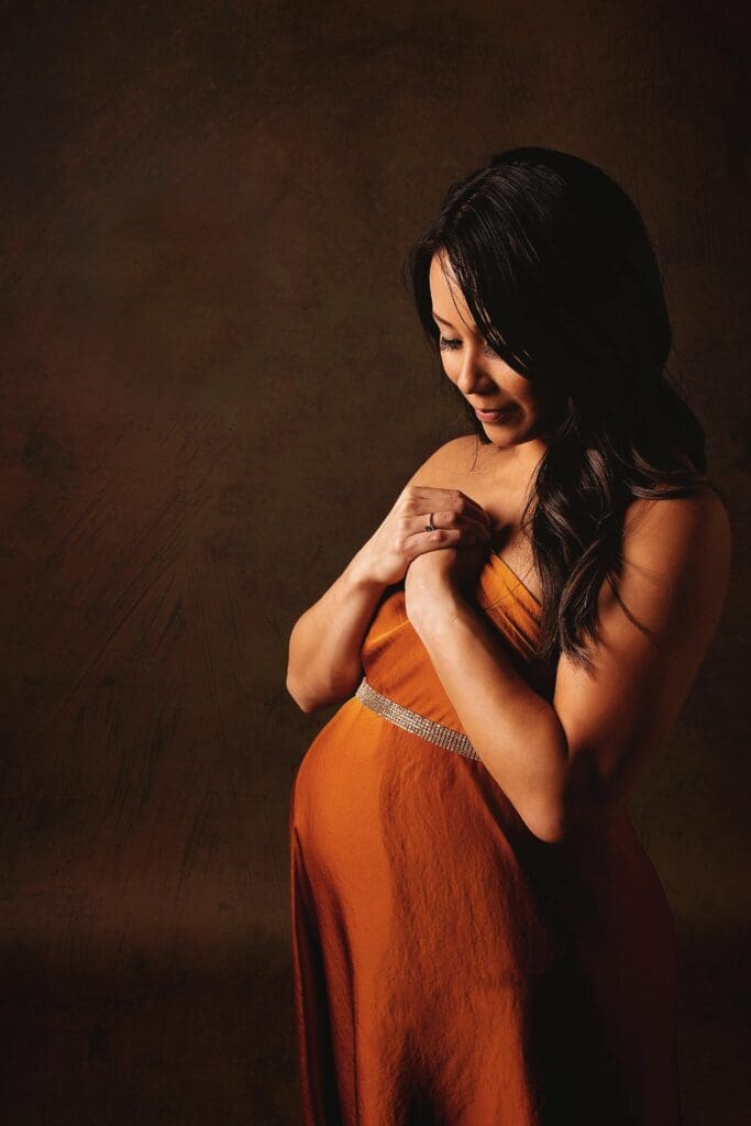 Fine art maternity portrait of an expectant mother in a rust-colored gown at a Nashville maternity photography studio, softly embracing her baby bump with dramatic studio lighting.