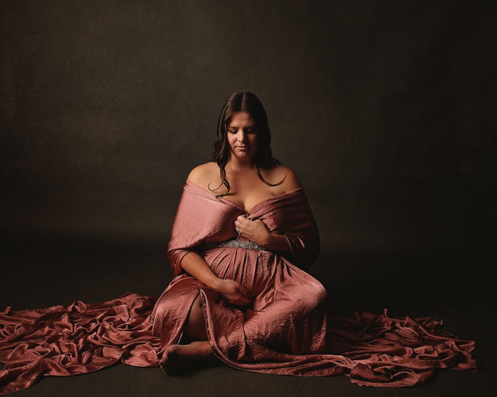 Expectant mother in a flowing rose gown sits peacefully with her hands on her baby bump during a fine art Nashville maternity photography session in a warm, dramatic studio.