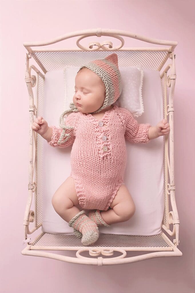 Sleeping newborn wearing a hand-knit pink romper and bonnet in a vintage metal bed during a fine art Nashville newborn photography session.