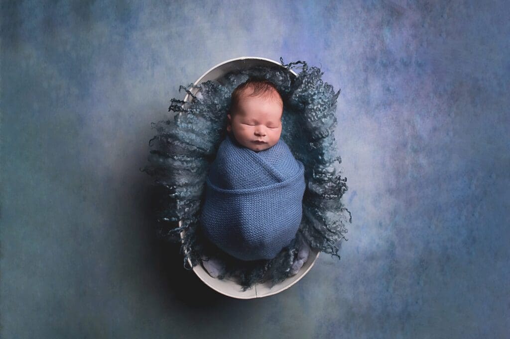 Sleeping newborn baby wrapped in a soft blue knit swaddle and nestled in a white bowl with textured blue layers during a fine art studio newborn photography session.