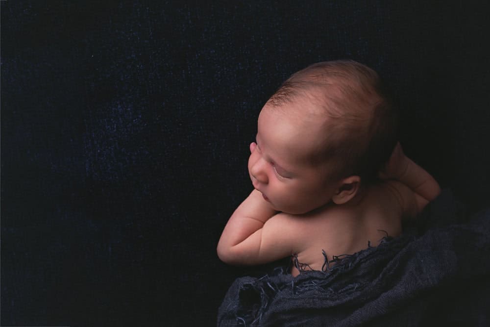 Sleeping newborn baby posed on their tummy with hands tucked beneath their head against a dark navy backdrop during a fine art studio portrait session.