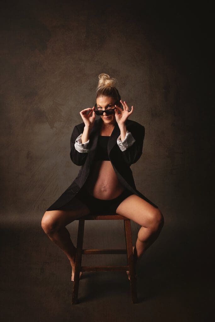 Confident expectant mother wearing a black blazer and sunglasses during a fine art Nashville maternity photography session, seated on a stool in a dramatic studio portrait.