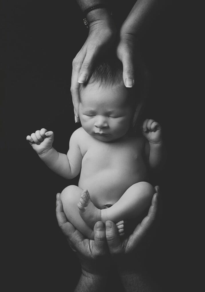 Black and white fine art portrait of a sleeping newborn cradled in their parents' hands against a dark background, highlighting the love and protection surrounding a new baby.