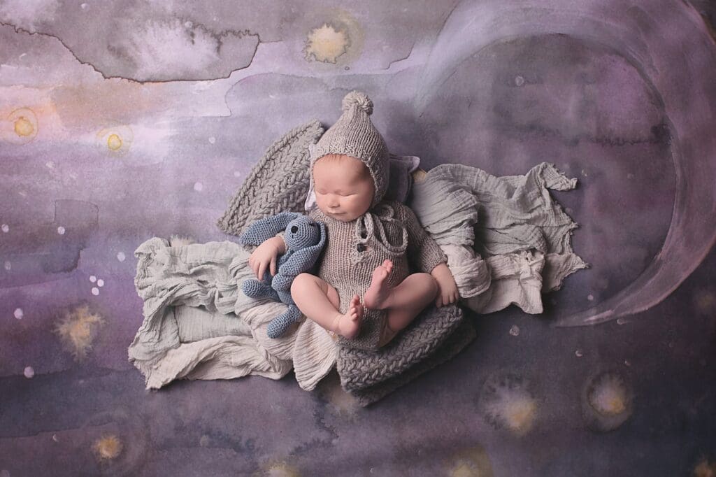 Fine art newborn portrait of a sleeping baby in a hand-knit outfit and bonnet, cuddling a knitted blue bunny on a dreamy moon backdrop during a Nashville newborn photography session.