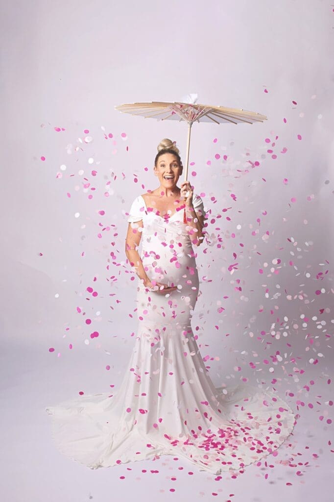 Joyful expectant mother in a flowing white gown holding a paper parasol as pink and white confetti falls during a Nashville maternity photography session and gender reveal.