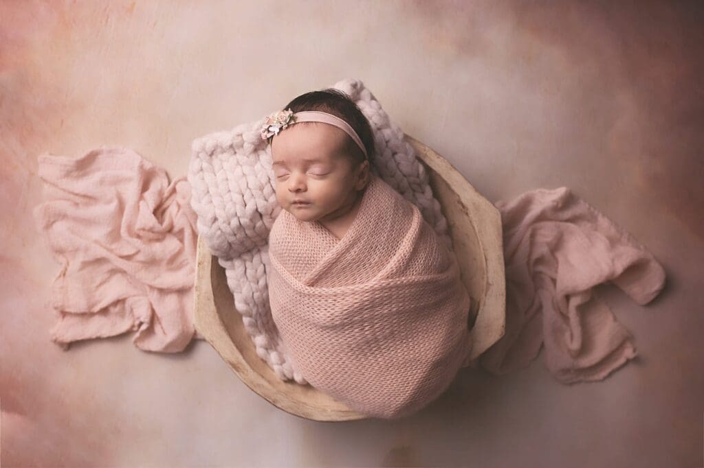 Sleeping newborn wrapped in a soft blush swaddle with a delicate floral headband, nestled in a wooden bowl during a fine art Nashville newborn photography session.