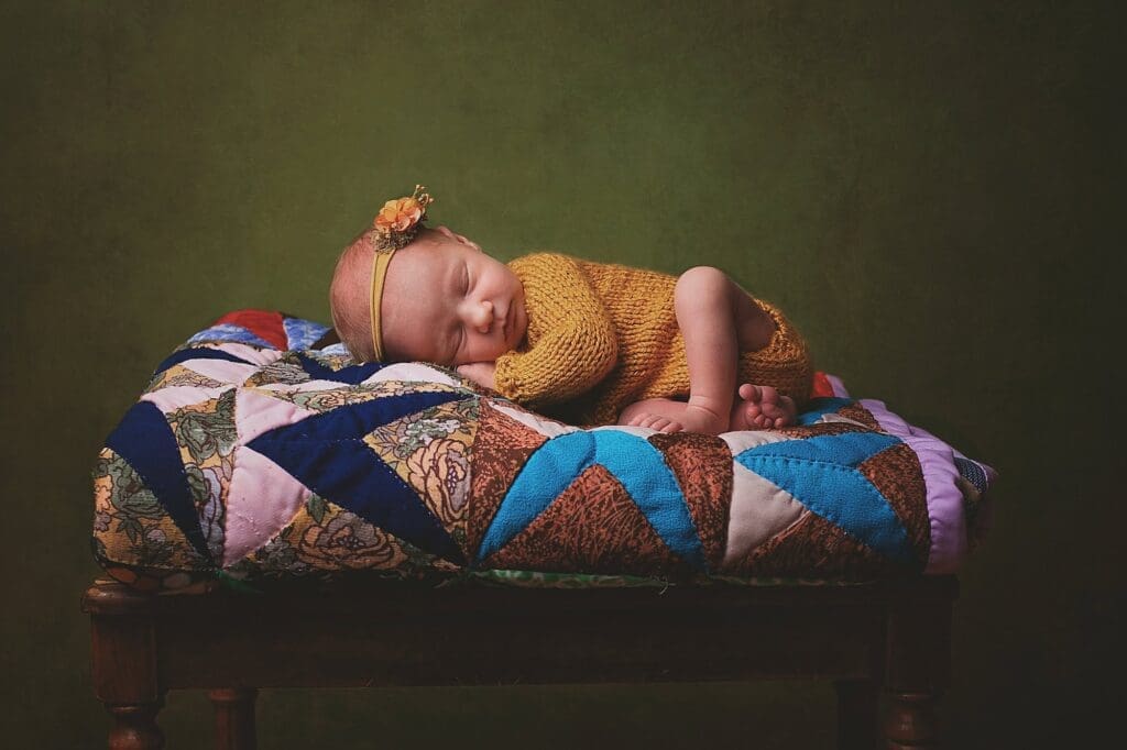 Sleeping newborn wearing a hand-knit mustard romper and floral headband, resting on a colorful vintage quilt during a fine art Nashville newborn photography session.