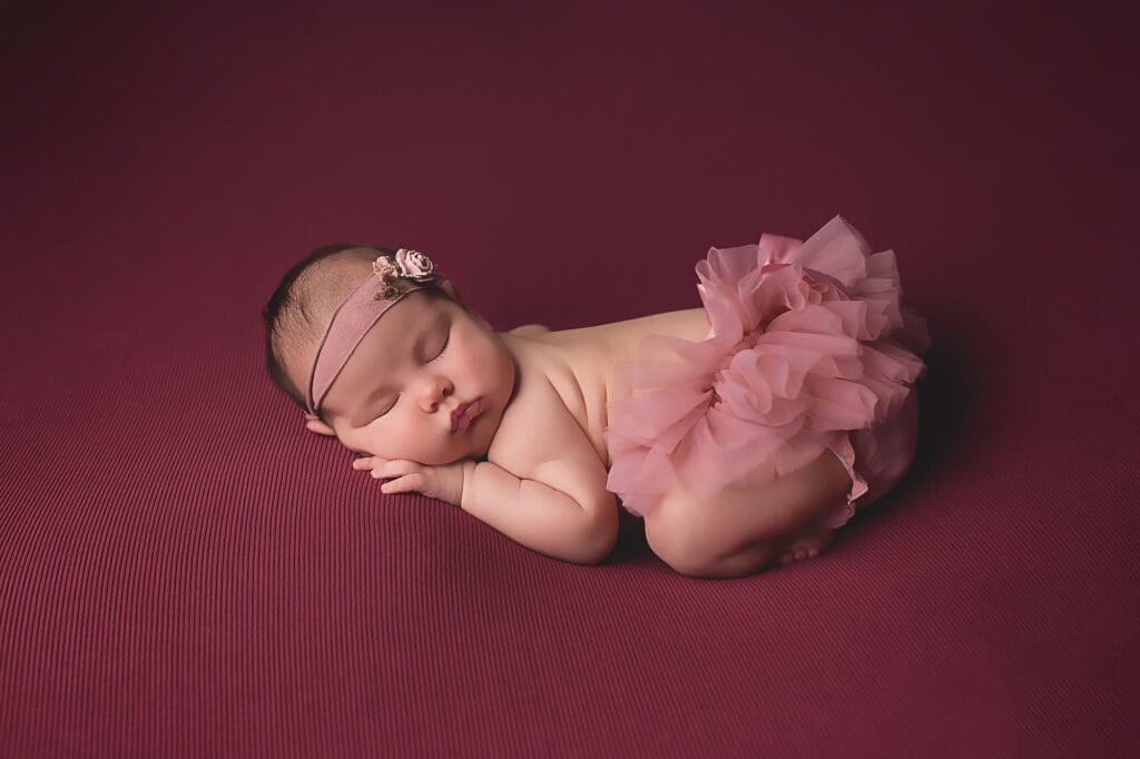 Sleeping newborn baby wearing a soft pink tutu and delicate floral headband, posed on a deep burgundy backdrop during a fine art studio newborn photography session.