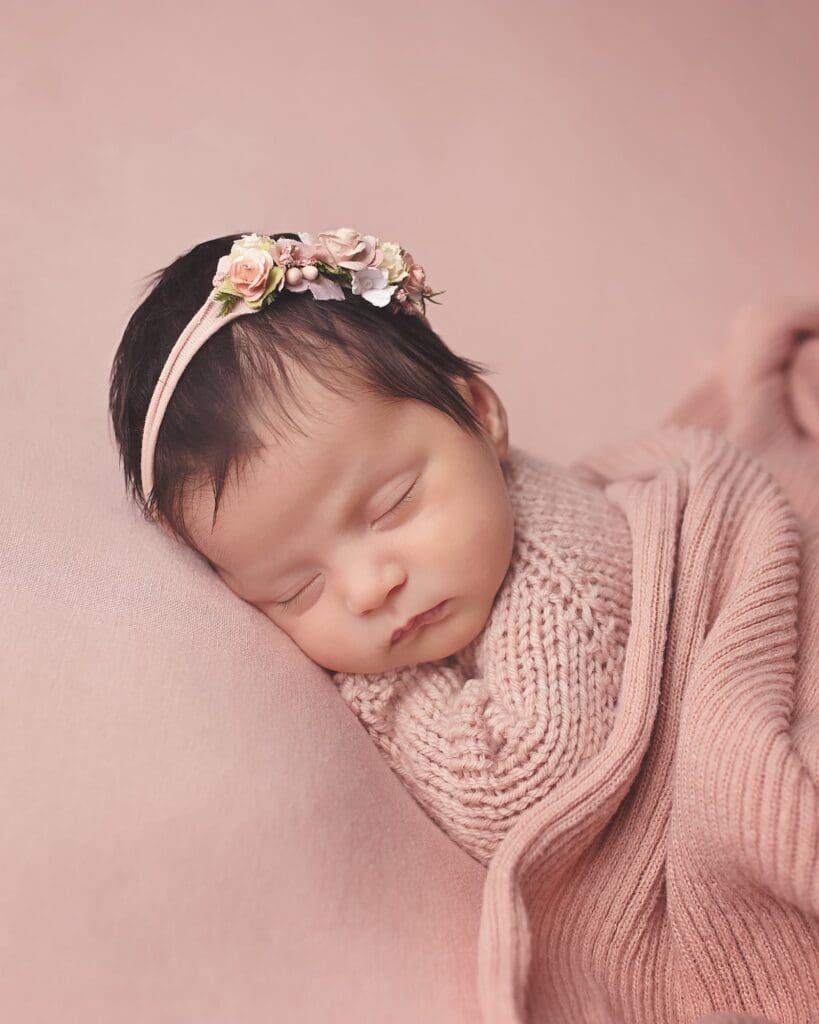Sleeping newborn baby wrapped in a soft blush pink knit blanket and sweater, wearing a delicate floral headband against a matching pink backdrop during a fine art studio portrait session.