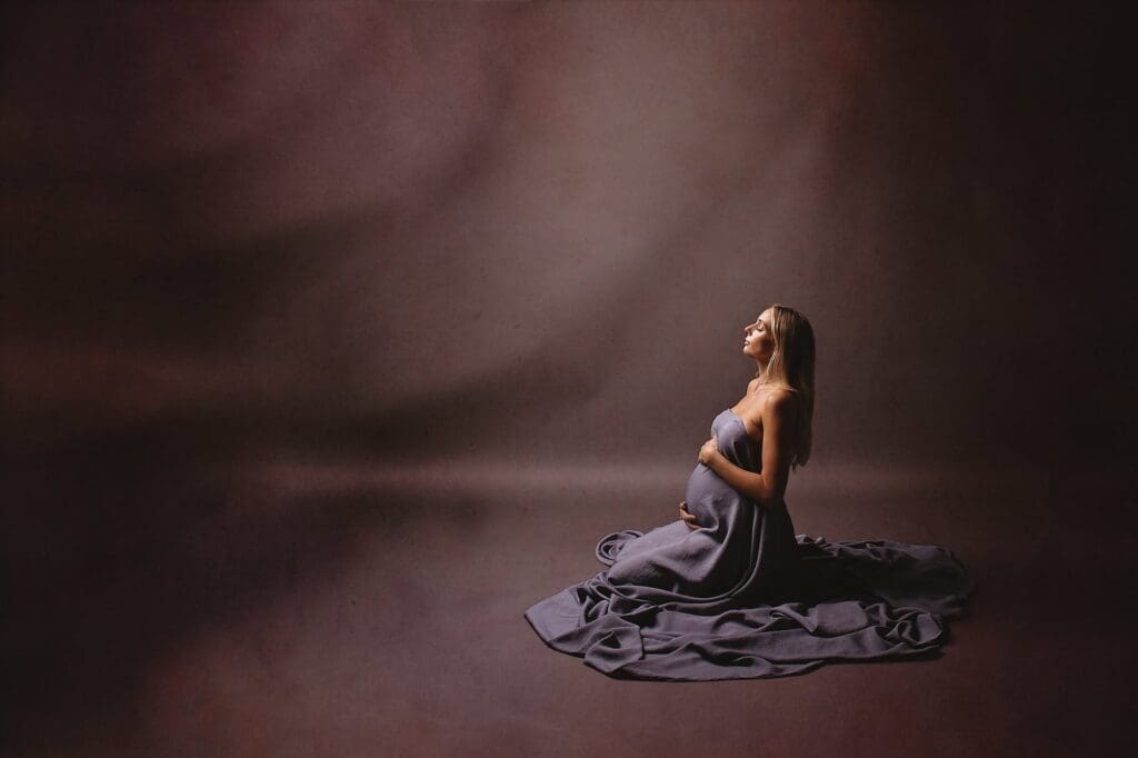 Peaceful fine art maternity portrait of an expectant mother seated in a flowing blue gown during a Nashville maternity photography session, gently holding her baby bump in dramatic studio lighting.