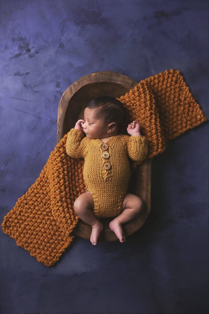 Sleeping newborn wearing a hand-knit mustard romper, resting in a wooden bowl with a chunky knit blanket during a Nashville newborn photography session.