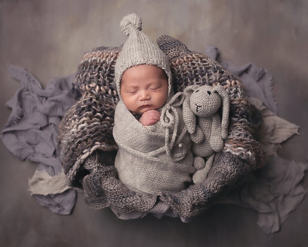 Sleeping newborn wrapped in a soft gray swaddle with a hand-knit bonnet, cuddling a knitted bunny during a fine art Nashville newborn photography session.