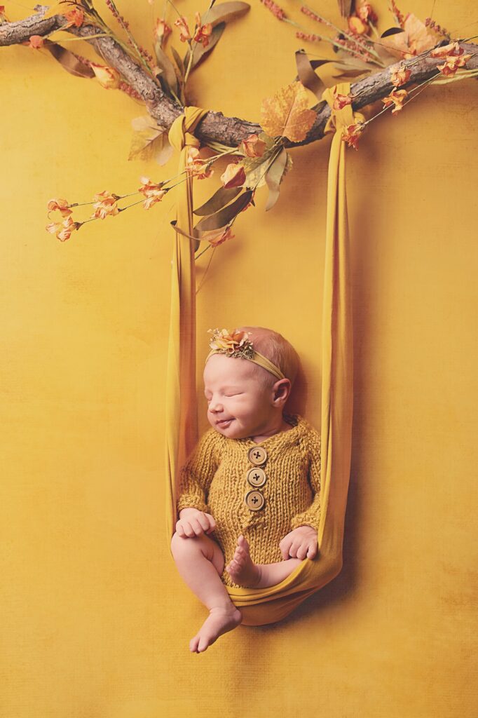 Sleeping newborn baby wearing a mustard knit outfit and floral headband, nestled in a hanging fabric swing beneath an autumn branch during a Nashville newborn photography session.