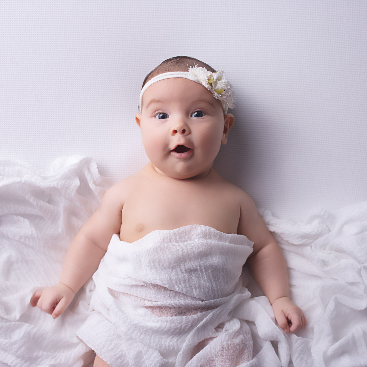 Smiling baby girl wrapped in a soft white swaddle, wearing a delicate white floral headband and looking toward the camera during a Nashville milestone photography session.