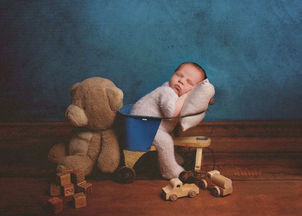 Sleeping newborn baby posed in a vintage blue wagon with a teddy bear, wooden toys, and alphabet blocks against a rich blue backdrop during a Nashville newborn photography session.