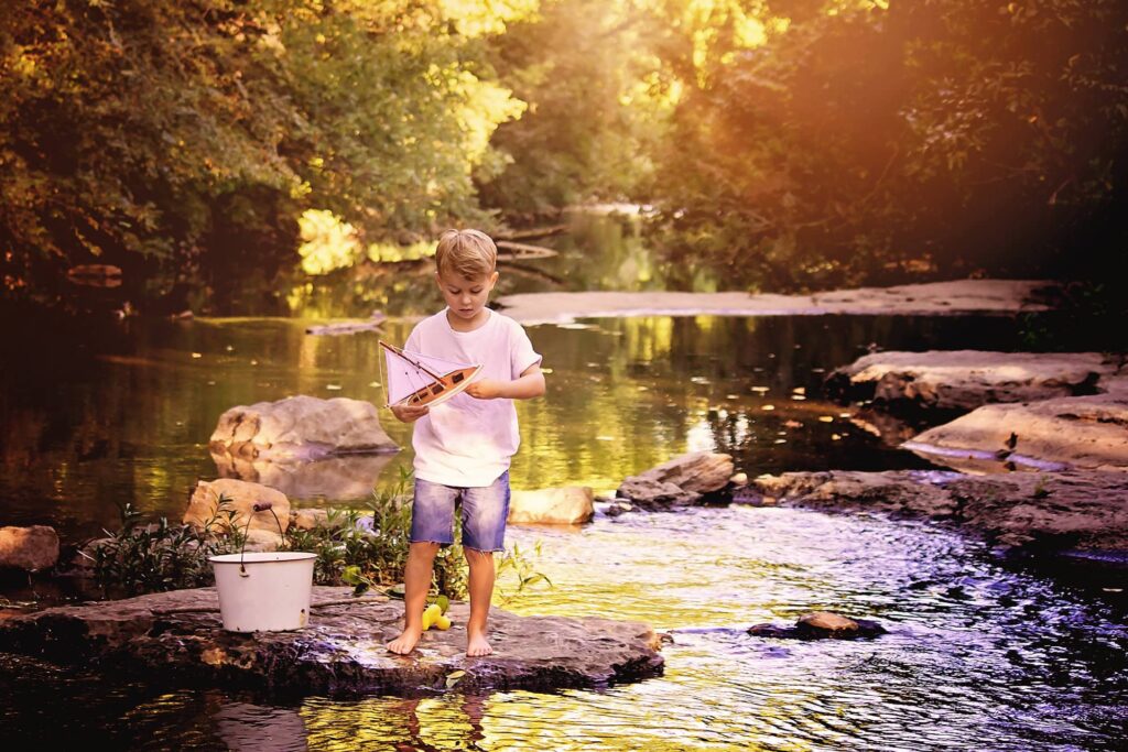 Young boy standing barefoot on rocks in a peaceful creek, holding a toy sailboat during a golden-hour Nashville child photography session.