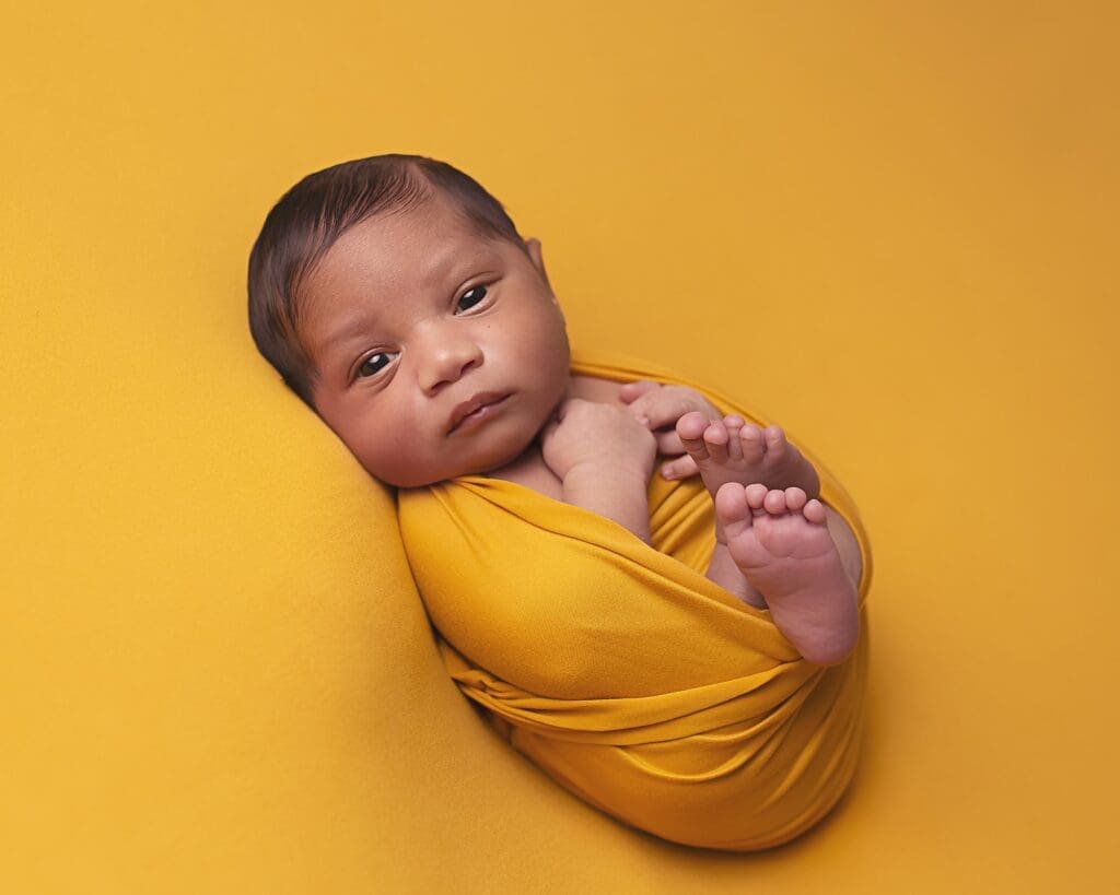 Awake newborn baby wrapped in a vibrant mustard yellow wrap, looking toward the camera.