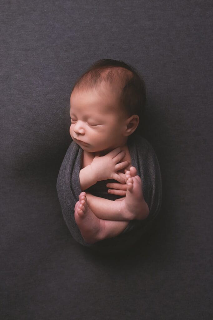Peacefully sleeping newborn baby wrapped in a charcoal gray wrap with tiny feet tucked in during a timeless Nashville newborn photography session.