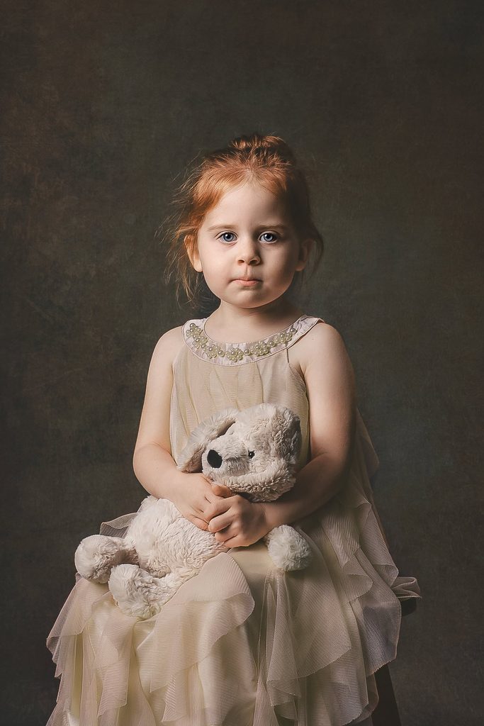 Fine art portrait of a young red-haired girl holding a favorite stuffed bunny against a dark painted backdrop.