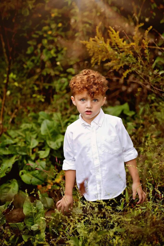 Young boy with curly red hair wearing a white shirt, standing among lush greenery during an outdoor Nashville child photography session.