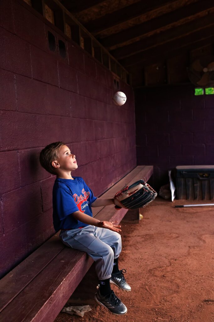 Young baseball player sitting in a dugout tossing a baseball into the air, captured during a Nashville childhood photography session.