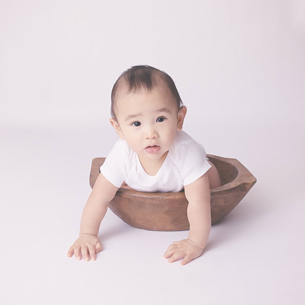 Baby leaning forward from a rustic wooden bowl against a soft neutral studio background.