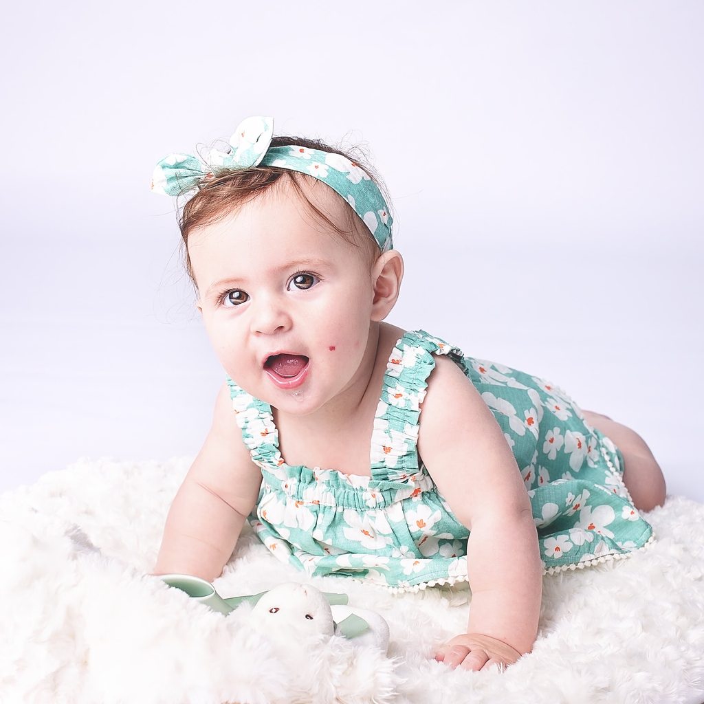 Baby girl in a mint floral romper and matching headband lying on a soft white blanket during a milestone portrait session.
