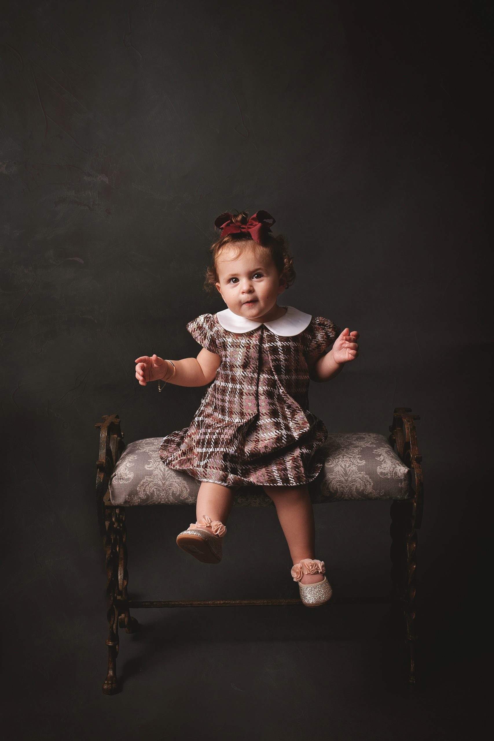 Toddler girl in a plaid dress with a burgundy hair bow sitting on a vintage upholstered bench against a dark fine art backdrop.