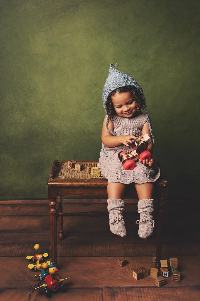 Young girl in a hand-knit dress and bonnet playing with vintage toys against a textured green backdrop for a Nashville child portrait.