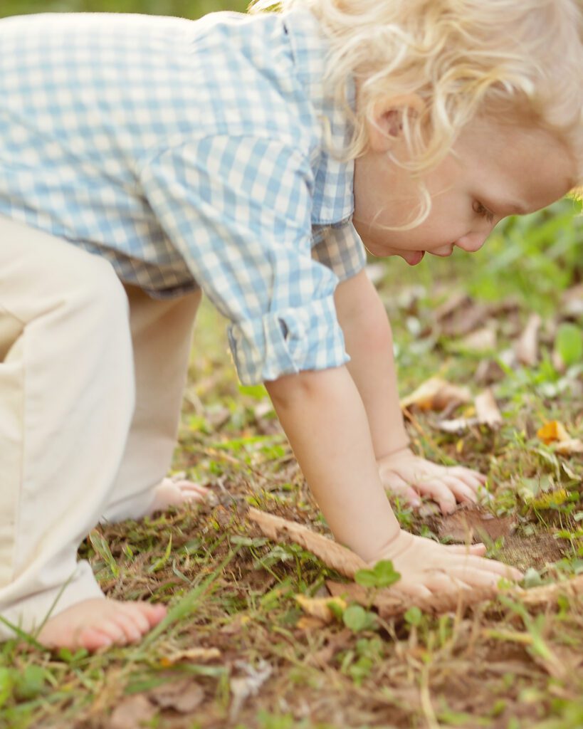 Curly-haired toddler exploring leaves and grass outdoors during a natural childhood portrait session in Nashville.