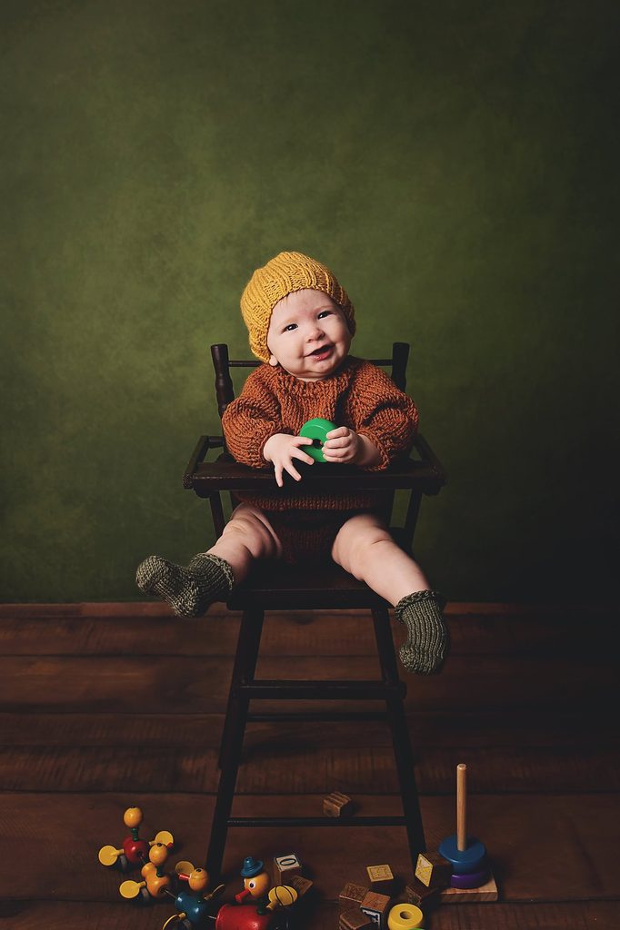 Smiling baby in a knitted outfit and mustard hat sitting in a vintage high chair with colorful toys.
