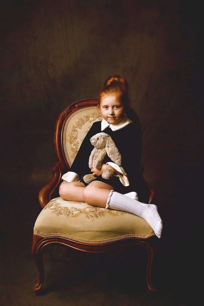 Young girl holding a stuffed bunny while seated on an antique chair in a dark fine art child portrait.