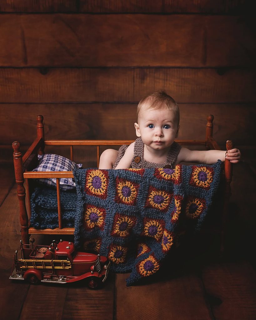 Baby boy sitting in a vintage wooden doll bed with a colorful crocheted blanket and antique toy fire truck.