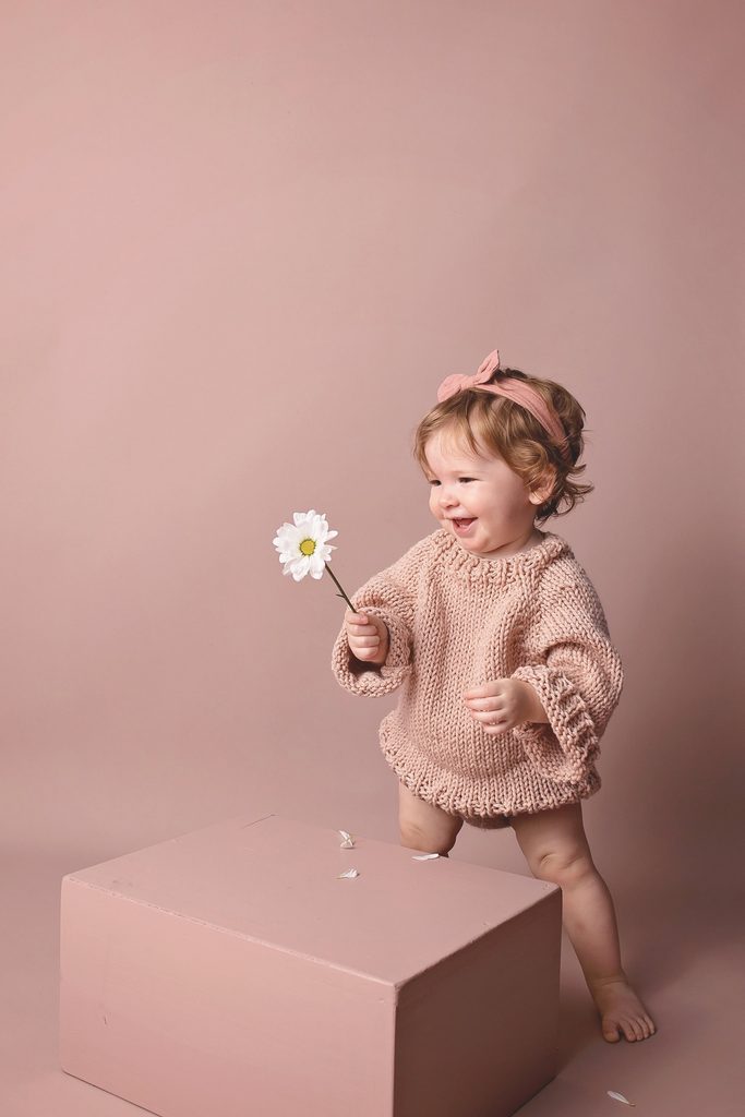 Smiling toddler girl in a hand-knit pink sweater holding a white daisy against a soft blush studio background.