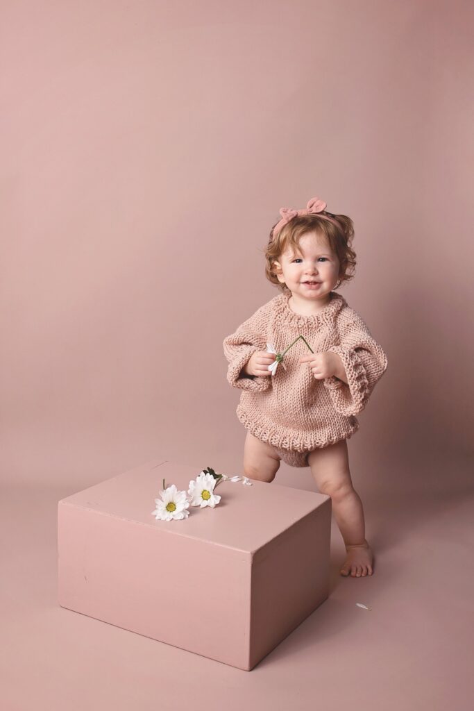 Smiling toddler girl in a hand-knit pink sweater holding daisies against a monochromatic blush backdrop by a Nashville child photographer.