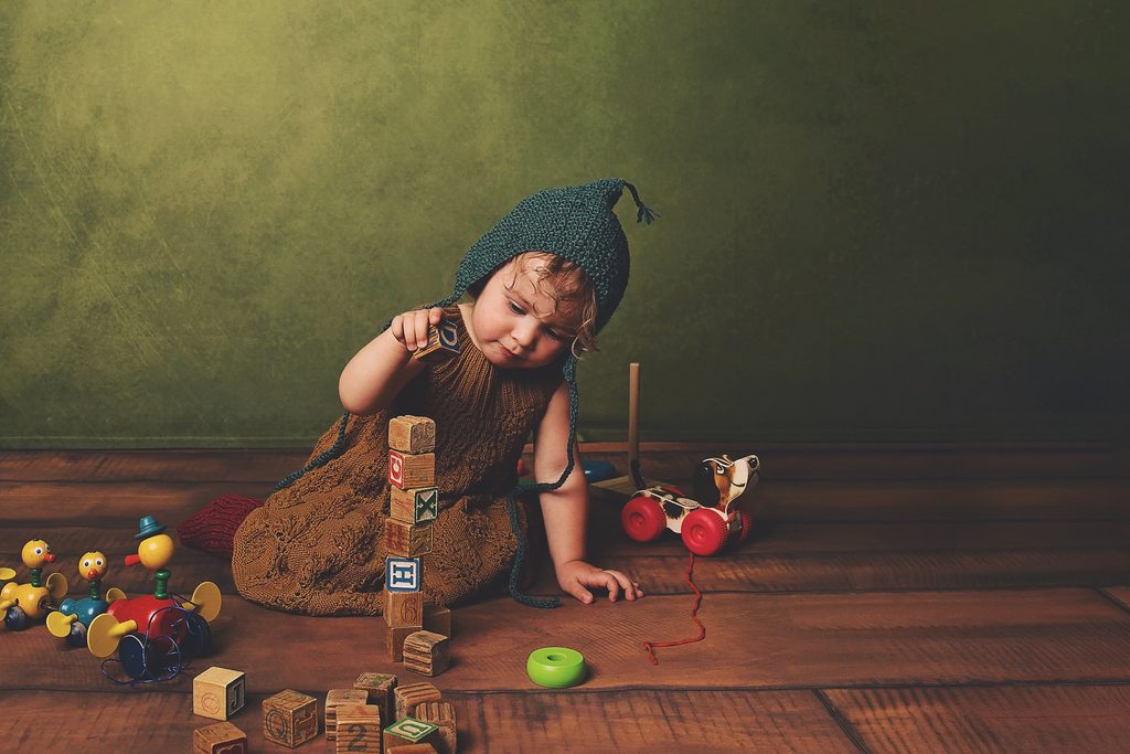 Toddler in a knitted bonnet and dress stacking vintage wooden blocks with colorful toys during a fine art Nashville child photography session.