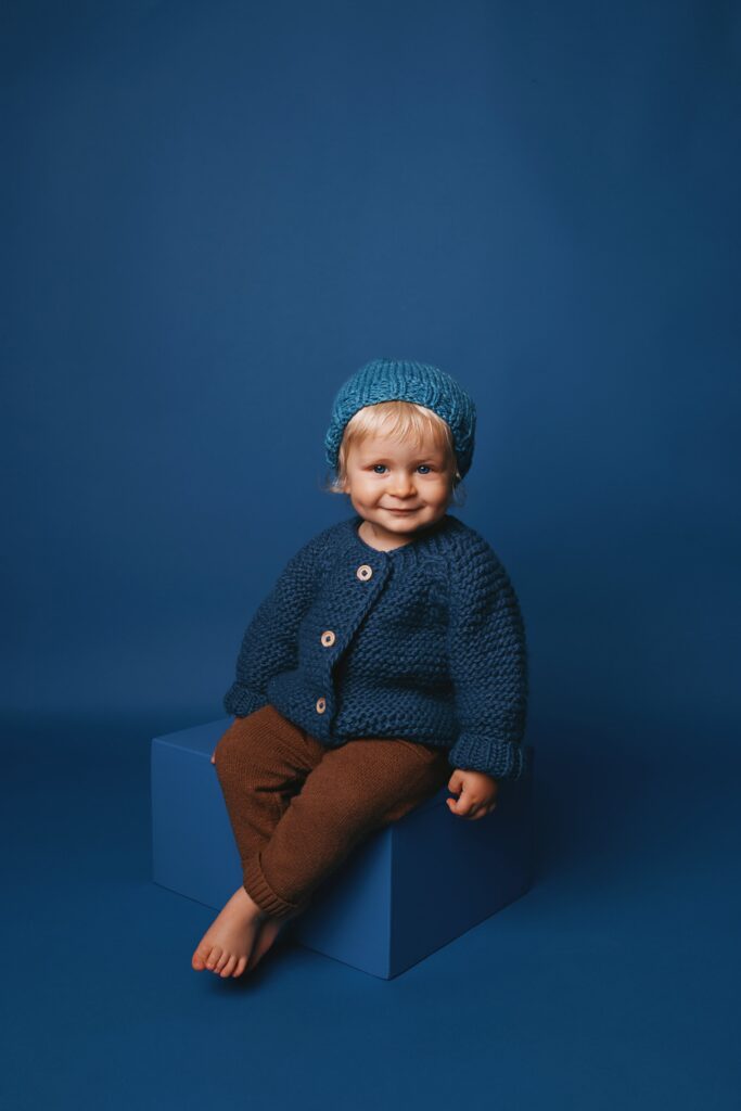 Blond toddler wearing a hand-knit blue sweater and hat seated against a monochromatic blue backdrop for a Nashville child portrait.