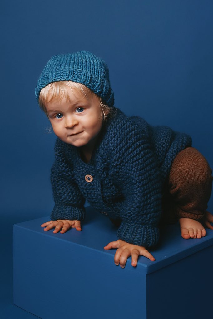 Blond toddler wearing a hand-knit blue sweater and hat crouching on a blue cube against a monochromatic blue backdrop for a Nashville child portrait.