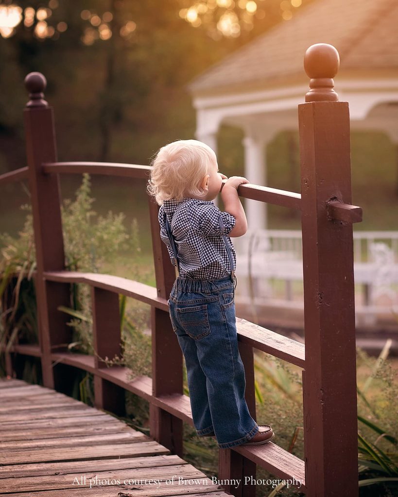 Blond toddler peeking over a wooden bridge railing in warm evening light during an outdoor Nashville child photography session.