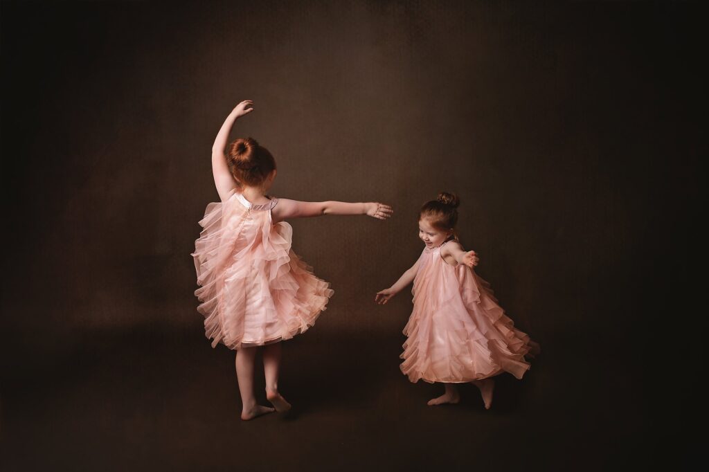 Two young sisters twirl barefoot in matching blush pink ruffled dresses against a dark brown studio backdrop during a fine art sibling portrait session with a Nashville children's photographer.