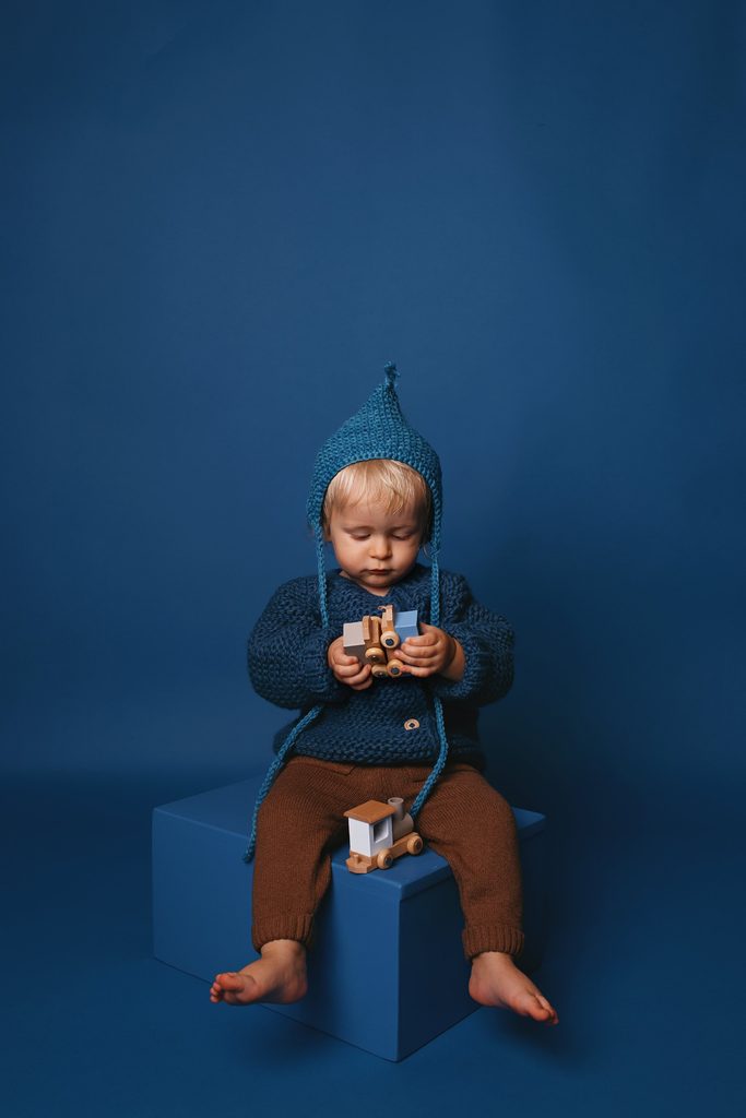 Blond toddler in a hand-knit blue outfit playing with wooden toy trains against a monochromatic blue backdrop by a Nashville child photographer.
