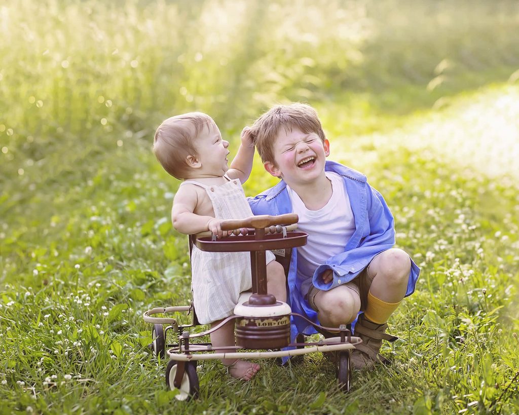 Baby and older brother laughing together beside a vintage tricycle in a sunlit field during a Nashville children’s photography session.